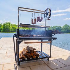 an outdoor grill with wood and sausages cooking on it's side by the water