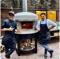 two men in aprons standing next to an outdoor pizza oven with wood on it