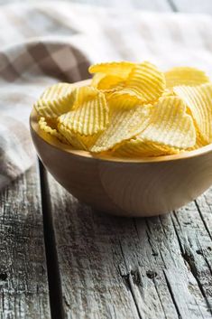 a wooden bowl filled with yellow potato chips on top of a wooden table next to a checkered cloth