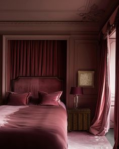 a bed with pink sheets and pillows in a bedroom next to a window covered by red curtains