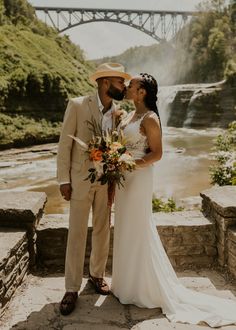 a bride and groom are standing in front of a waterfall with a bridge behind them