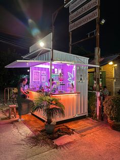 a woman standing in front of a food stand at night with purple lights on it