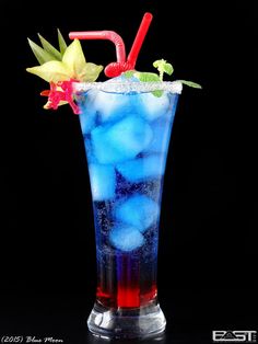 a tall glass filled with blue liquid and ice next to a flower on the side