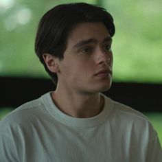 a young man in a white t - shirt looks off into the distance while sitting down