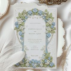 an ornate wedding card with blue flowers and greenery on the front is surrounded by feathers