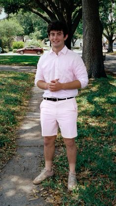 a man in pink shirt and white shorts standing on sidewalk next to tree with grass