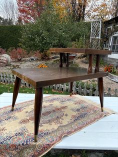 a wooden table sitting on top of a rug next to a building and trees in the background