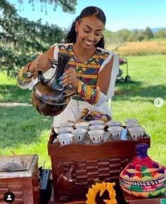 a woman is pouring tea into cups at an outdoor picnic table with other items on the grass