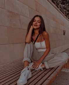 a woman sitting on a wooden bench next to a stone wall and wearing white sneakers