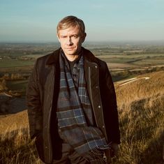 a man standing on top of a grass covered hill next to a field with rolling hills in the background