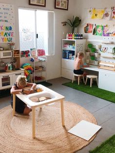 a child sitting in a playroom with toys and books on the floor, while another child sits at a table