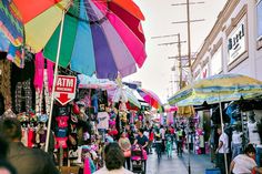 many people are walking through an open market area with umbrellas and other items on display