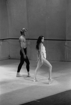 two people standing in an empty room with one person wearing tight pants and the other holding a ballet shoe