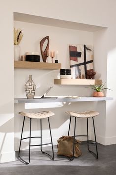 two stools sitting in front of a white counter with shelves above it and a vase on the wall