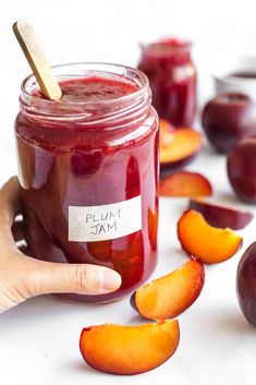 plum jam in a glass jar with peach slices around it and a wooden stick sticking out of the jar