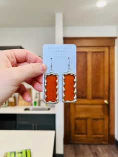 a person holding up two earrings in front of a kitchen counter with wooden doors and cabinets