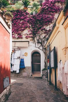 an alley way with clothes hanging on the wall and purple flowers growing over the doorway