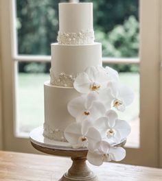 a three tiered cake with white flowers on the top and bottom is sitting on a table in front of a window