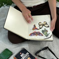 a woman sitting on the ground holding an open book with butterflies painted on it and other items around her