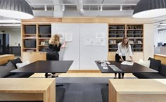 two women standing in an office setting with desks and chairs, one woman is writing on a whiteboard