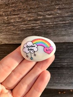 a hand holding a white rock with a rainbow baby on it and a pink heart in the middle