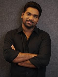 a man with his arms crossed standing in front of a gray wall wearing a black shirt