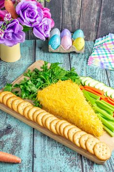 an assortment of vegetables and crackers on a wooden table next to easter eggs, carrots and flowers