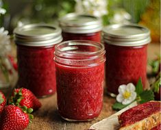 four jars filled with strawberry jam sitting on top of a wooden table next to sliced strawberries