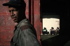 a man standing in front of a red building with people sitting on the sidewalk behind him