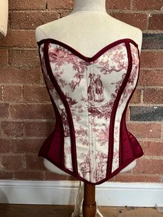 Overbust corset in burgundy satin with front panels in cotton printed with fabulous skeleton toile du jouy fabric.   The corset is made with a strength layer of Coutil and is boned with a mixture of flat and spiral steel bones, it is back lacing only with an unboned modesty panel.   The corset measures 27" at the waist and so would probably best suit natural waists in the region of 30-33"  Fully laced the corset measures   Bust 38"  Waist 27"  Upper hip 37" Length at front centre 14" Red Fitted Underbust Corset Dress, Red Underbust Corset Dress, Fitted Red Bodice With Corset Back, Red Fitted Corset With Boning, Red Corset With Boning, Fitted Red Corset With Boned Bodice, Red Fitted Bodice Corset With Boning, Red Overbust Corset Dress With Boning, Red Overbust Bodice With Boning