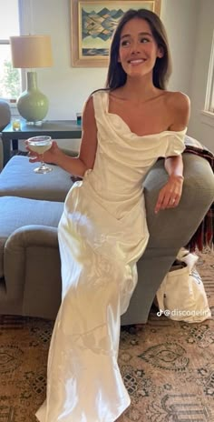 a woman in a white dress sitting on a couch holding a wine glass and smiling