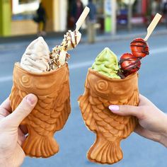 two people holding up ice cream cones with strawberries and cherries in them on the street