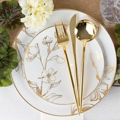 a white plate topped with gold colored utensils next to green leaves and flowers