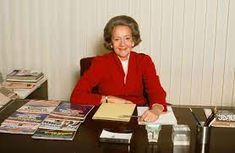 an older woman sitting at a desk with books and papers on it, smiling for the camera