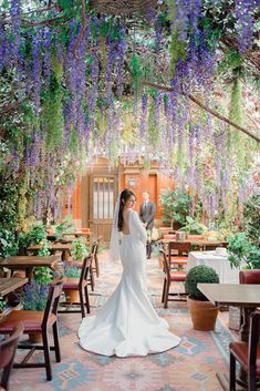 a bride and groom standing in the middle of a room with purple flowers hanging from the ceiling