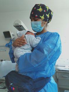 a woman holding a baby in a hospital room wearing a face mask and blue gown