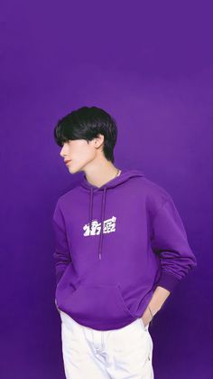 a male in a purple hoodie and white pants standing against a purple wall with his hands on his hips