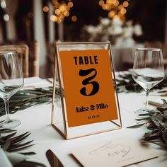 an orange table number sign sitting on top of a table next to wine glasses and napkins