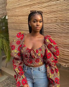 a woman with braids wearing a red top and jeans standing in front of a bamboo wall