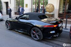 a black sports car parked in front of a building with people standing around it and two men walking on the sidewalk