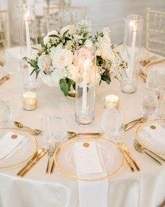 a table set with white and gold place settings, candles and flowers in vases