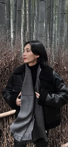 a woman standing in front of bamboo trees wearing a black jacket and scarf with her hands on her hips