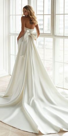 a woman in a white wedding dress with a bow on her back, looking out the window