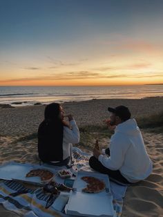 two people are sitting on the beach eating pizza and drinking wine while the sun sets