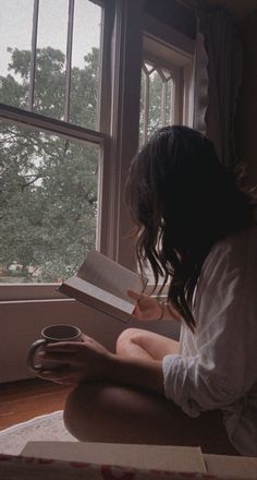 a woman sitting on the floor reading a book in front of a window with trees outside