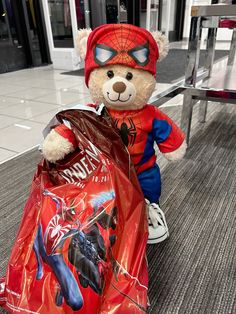 a teddy bear sitting on the ground with a bag in it's lap and wearing a spiderman costume