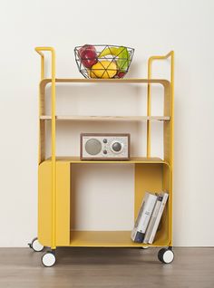 a yellow cart with books and a basket on top is shown in front of a white wall