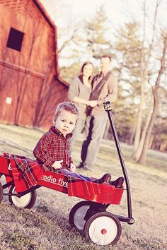 a little boy sitting in a red wagon with his parents standing behind him on the grass