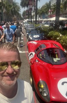 a man wearing sunglasses standing next to a red race car in front of other cars