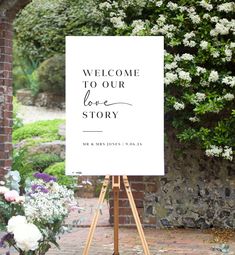 a welcome sign stands in front of a brick wall with white flowers and greenery